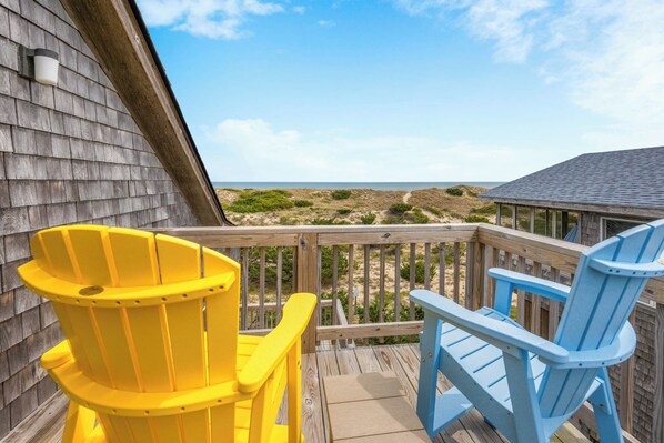A wooden deck with two Adirondack chairs, one yellow and one blue, overlooking a beach. - Oceanfront Dog Friendly Home with Easy Beach Access, Grill, and Smart TVs (Avon)