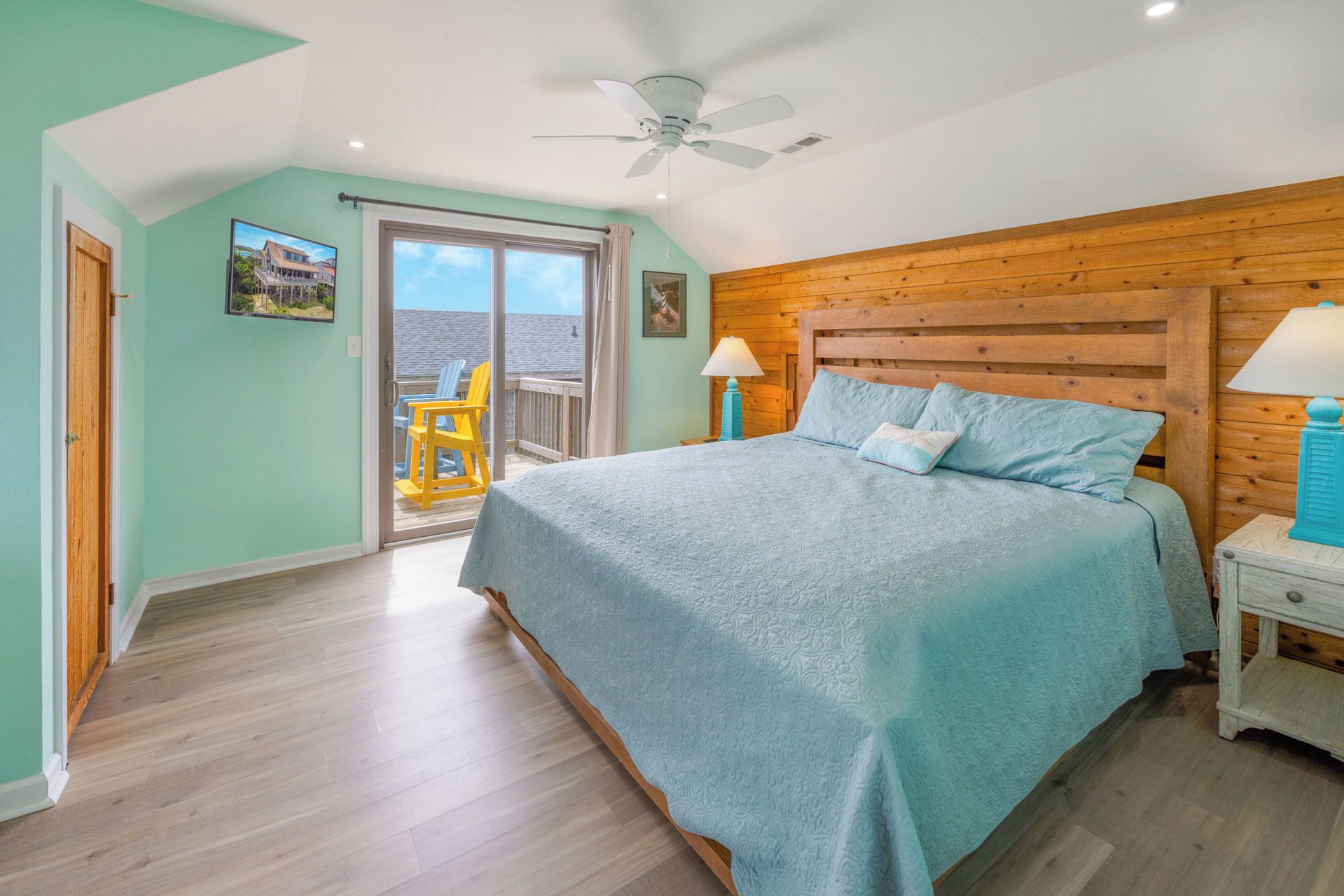 A bedroom with a large bed, wooden headboard, two bedside lamps, a ceiling fan, and a balcony with chairs.