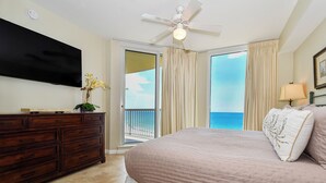 A bedroom with a bed, a dresser, a TV, a ceiling fan, and a balcony with a view of the ocean.