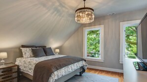 Une chambre à coucher avec un lit, des tables de chevet, un lustre, une fenêtre donnant sur un paysage verdoyant et un bureau avec un ordinateur portable.
