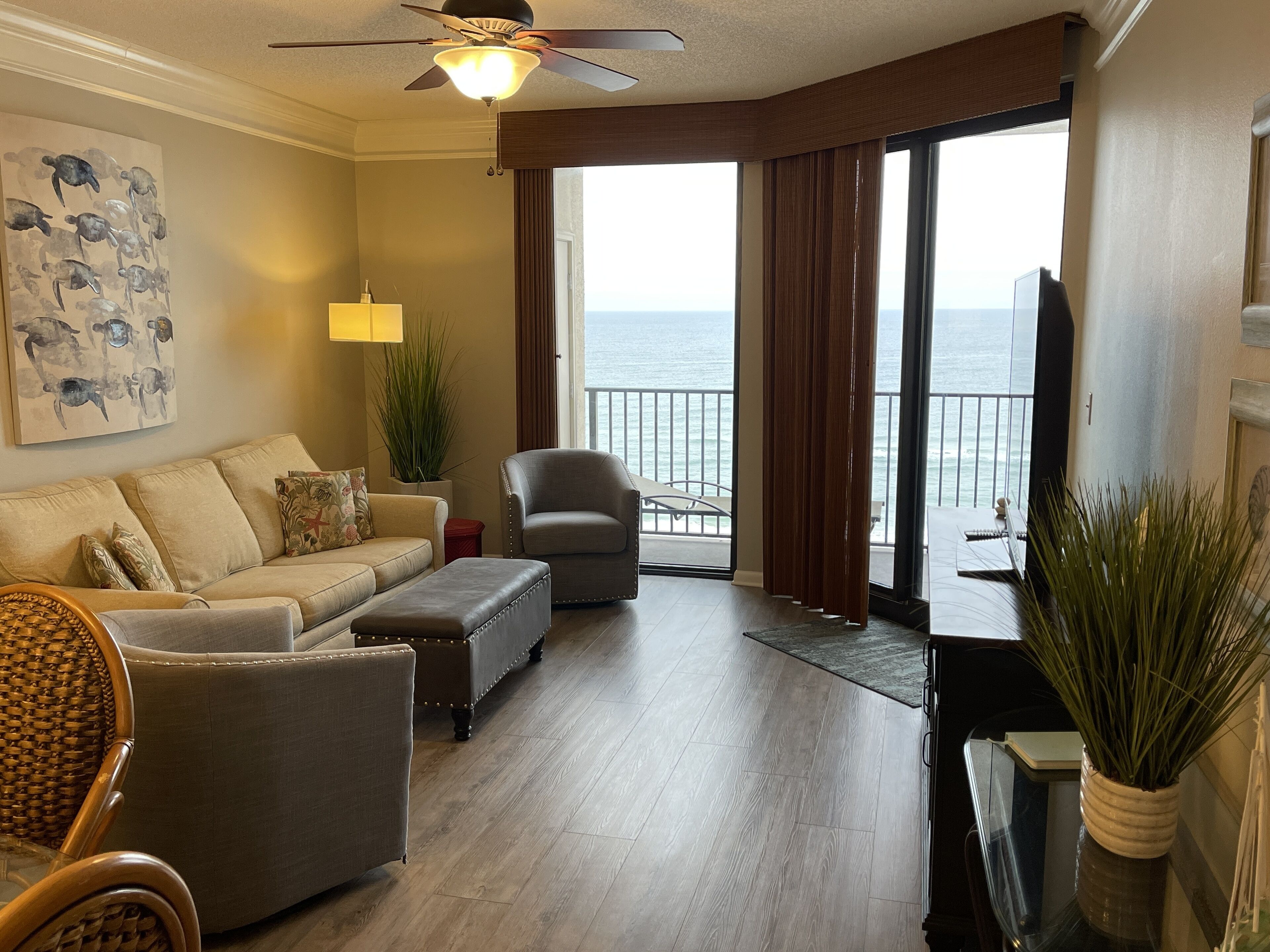 A living room with a sofa, armchair, and ottoman, a ceiling fan, a potted plant, and a view of the ocean through large windows.
