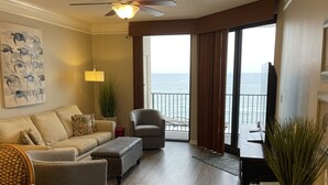 Un salon comprenant un canapé, un fauteuil et un pouf, un ventilateur de plafond, une plante en pot et une vue sur l’océan par de grandes fenêtres.