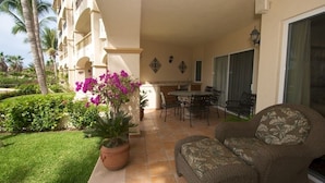 A balcony with a dining table, chairs, and a sofa. There are potted plants and a palm tree in the background.