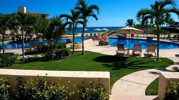 A resort pool area with lounge chairs and umbrellas, surrounded by palm trees and a view of the ocean.