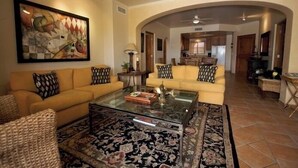 A spacious living room with a yellow sofa, a glass coffee table, and a large abstract painting.