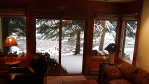 Ein gemütliches Wohnzimmer mit Kamin, Sofa, Couchtisch und Lampe. Große Fenster bieten Blick auf eine verschneite Landschaft mit Bäumen.