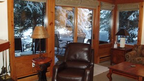 Ein gemütliches Wohnzimmer mit Ledersessel, Couchtisch aus Holz und einer Lampe. Große Fenster bieten Blick auf eine verschneite Landschaft.