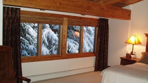 Ein Schlafzimmer mit Holzboden, einem großen Fenster mit Vorhängen, einem Nachttisch mit Lampe und einem Bett mit weißer Bettdecke.