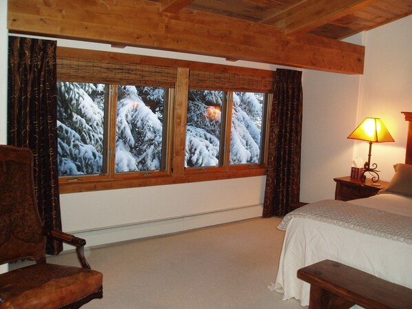 A bedroom with a wooden ceiling, a large window with curtains, a bedside table with a lamp, and a bed with a white comforter.