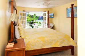 A bedroom with a bed, a nightstand, a lamp, a ceiling fan, a window with a view of greenery, and a painting on the wall. - Location Location Location+Top 1% in guest service on Big Island Oceanfront Home (Kailua-Kona)