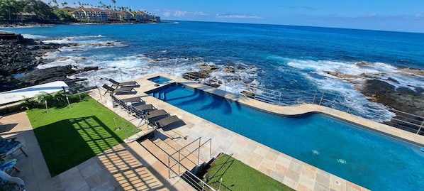 A pool with a view of the ocean, surrounded by lounge chairs and a grassy area.
