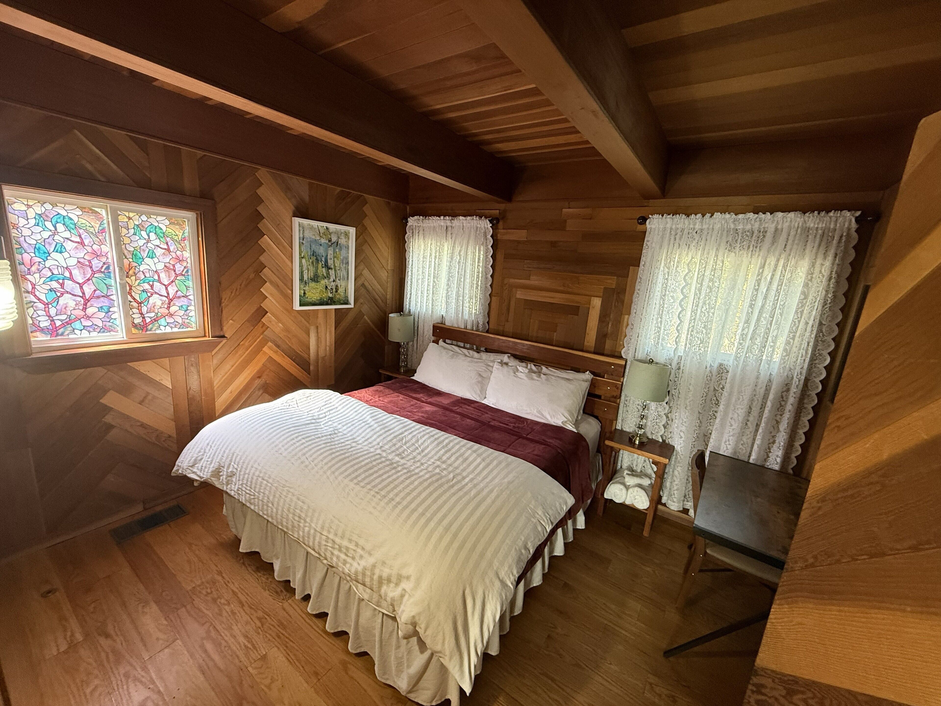 Une chambre à coucher aux murs lambrissés, comprenant un lit, une table de chevet, un tableau et une fenêtre ornée de vitraux.
