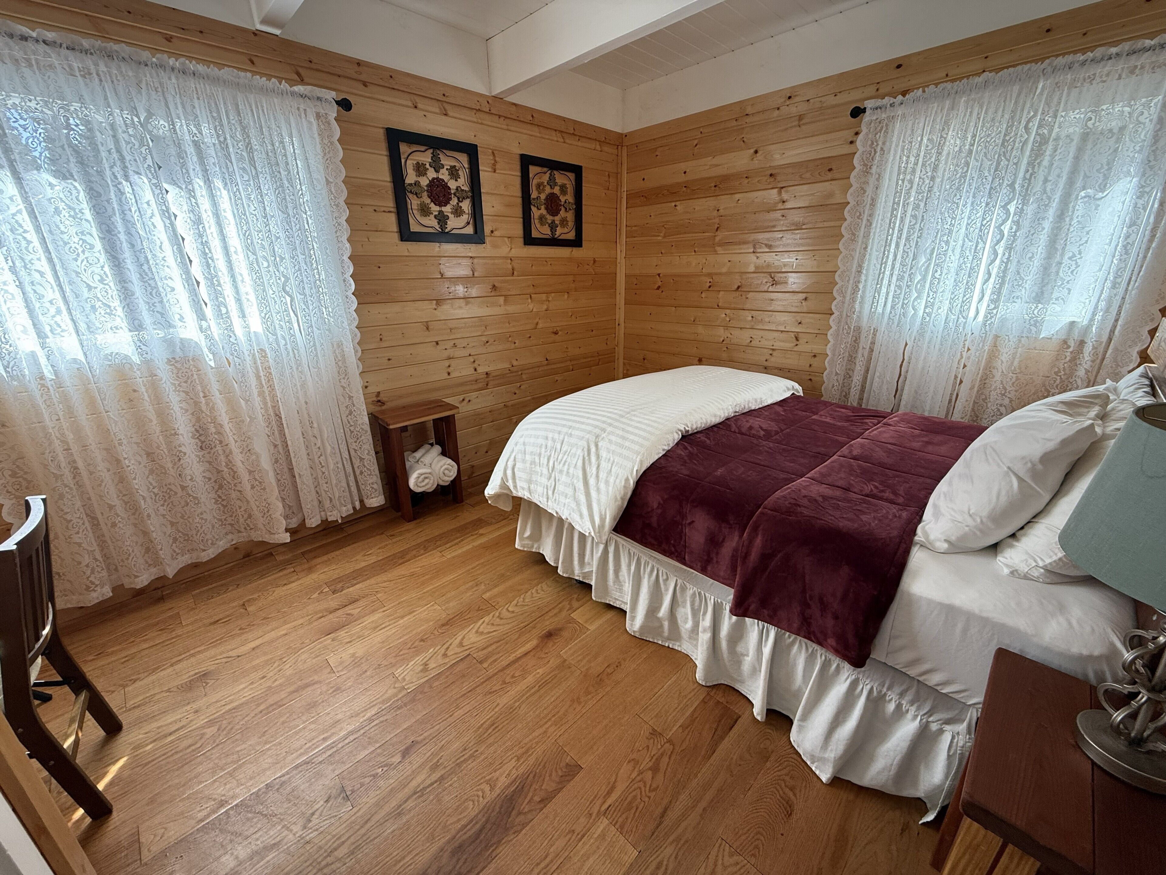 A wooden-paneled room with a bed, two framed floral wall art pieces, and a small wooden stool with towels.
