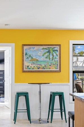 Une pièce avec un mur jaune, un tableau encadré représentant une scène de plage et deux tabourets verts.