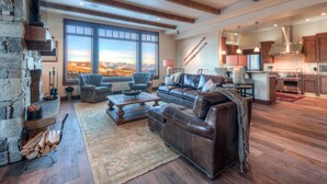 A spacious living area with a large window offering a mountain view, a fireplace, and a modern kitchen. - Deep Forest Retreat | Big Sky Ski-In/Out Luxury for 10 (Big Sky)