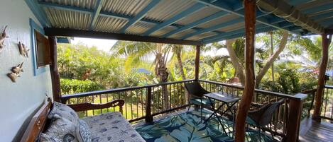 A covered outdoor seating area with a wooden bench and a table, surrounded by lush tropical vegetation.