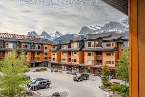 Un hotel de varios pisos con el paisaje de una montaña al fondo, que cuenta con un estacionamiento con vehículos y un pequeño espacio exterior para sentarse.