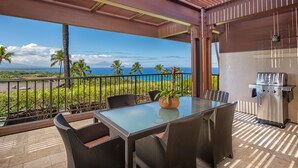 A covered outdoor dining area with a glass table, wicker chairs, a grill, and a view of the ocean. - Mauna Kea Villa, Walking Distance to the Beach and Hotel - Beyond Perfection! (Kamuela)