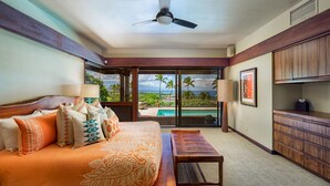 A hotel room with a bed, a desk, a view of a pool, and a ceiling fan. - Mauna Kea Villa, Walking Distance to the Beach and Hotel - Beyond Perfection! (Kamuela)
