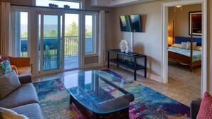 Un salon spacieux avec une table basse en verre, un canapé et un tapis coloré. Un téléviseur est fixé au mur et un balcon offre une vue sur l’extérieur.