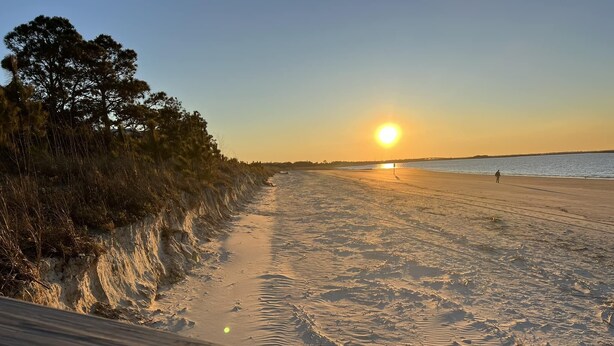 Une plage avec une promenade en bois menant à l’eau, un coucher de soleil en arrière-plan et une personne qui marche sur le sable.