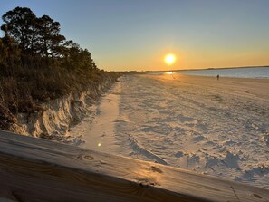 En strand med en träpromenad som leder ner till vattnet, en solnedgång i bakgrunden och en person som går på sanden.