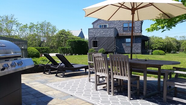 An outdoor patio with a grill, lounge chairs, and a table under a large umbrella. - Beautiful home with private heated pool, basketball and large outdoor space. (Nantucket)