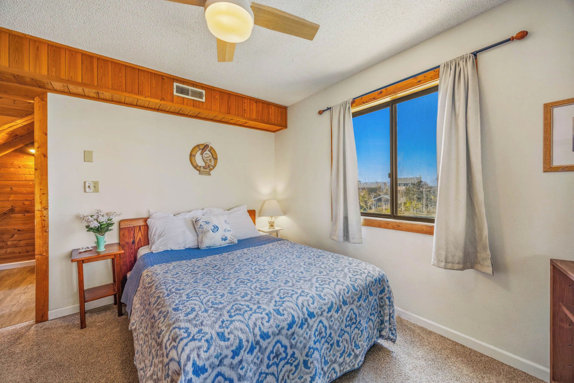 Un dormitorio con cama, mesita de noche, lámpara, ventana con cortinas, ventilador de techo y un panel de pared de madera.