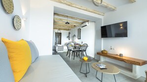 A modern living room with a sofa, a dining area with chairs, a TV mounted on the wall, and a wooden shelf. - Aelia Suites Milos (Milos)