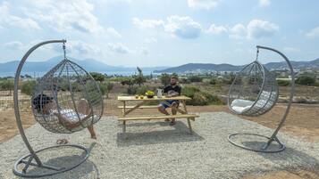 A picnic setup with a table and two hanging chairs, a person sitting on the table, and a scenic view of mountains and a body of water in the background.