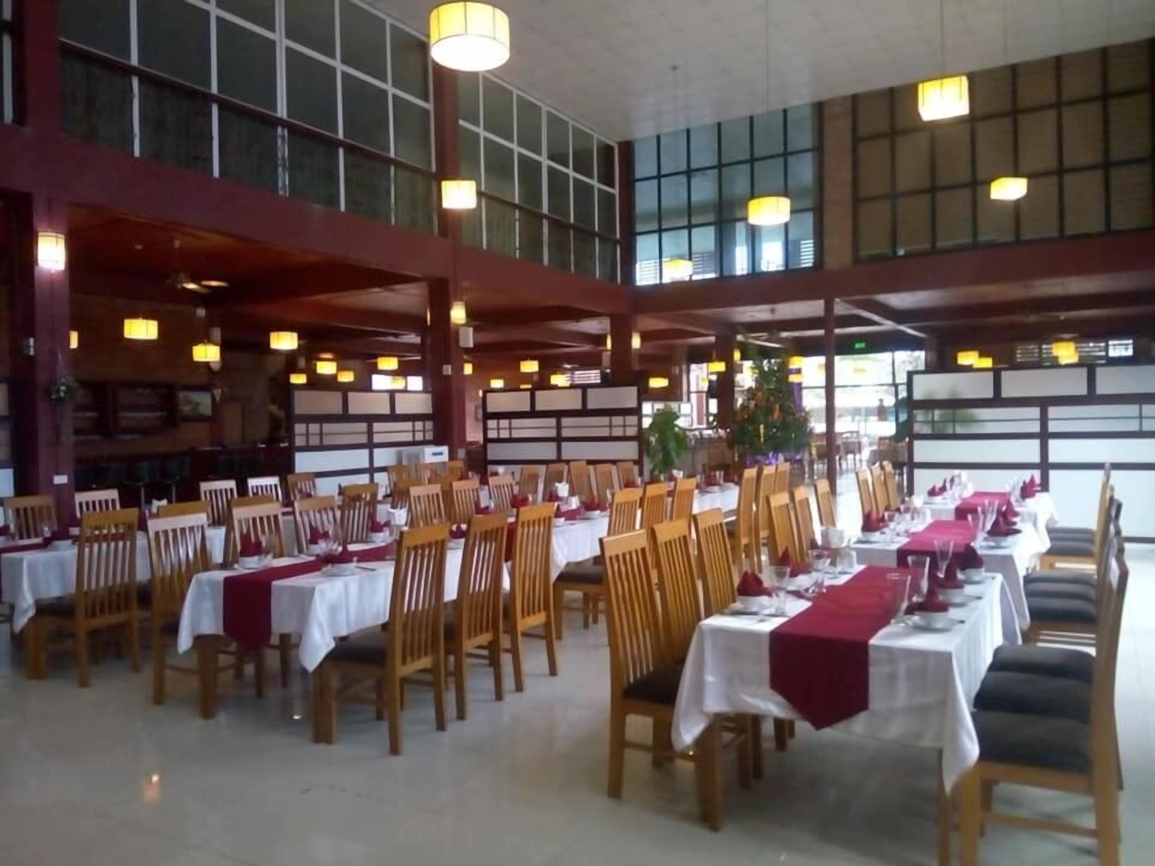 Ein geräumiger Speisesaal mit mehreren Tischen und Stühlen, großen Fenstern und hängenden Leuchten.