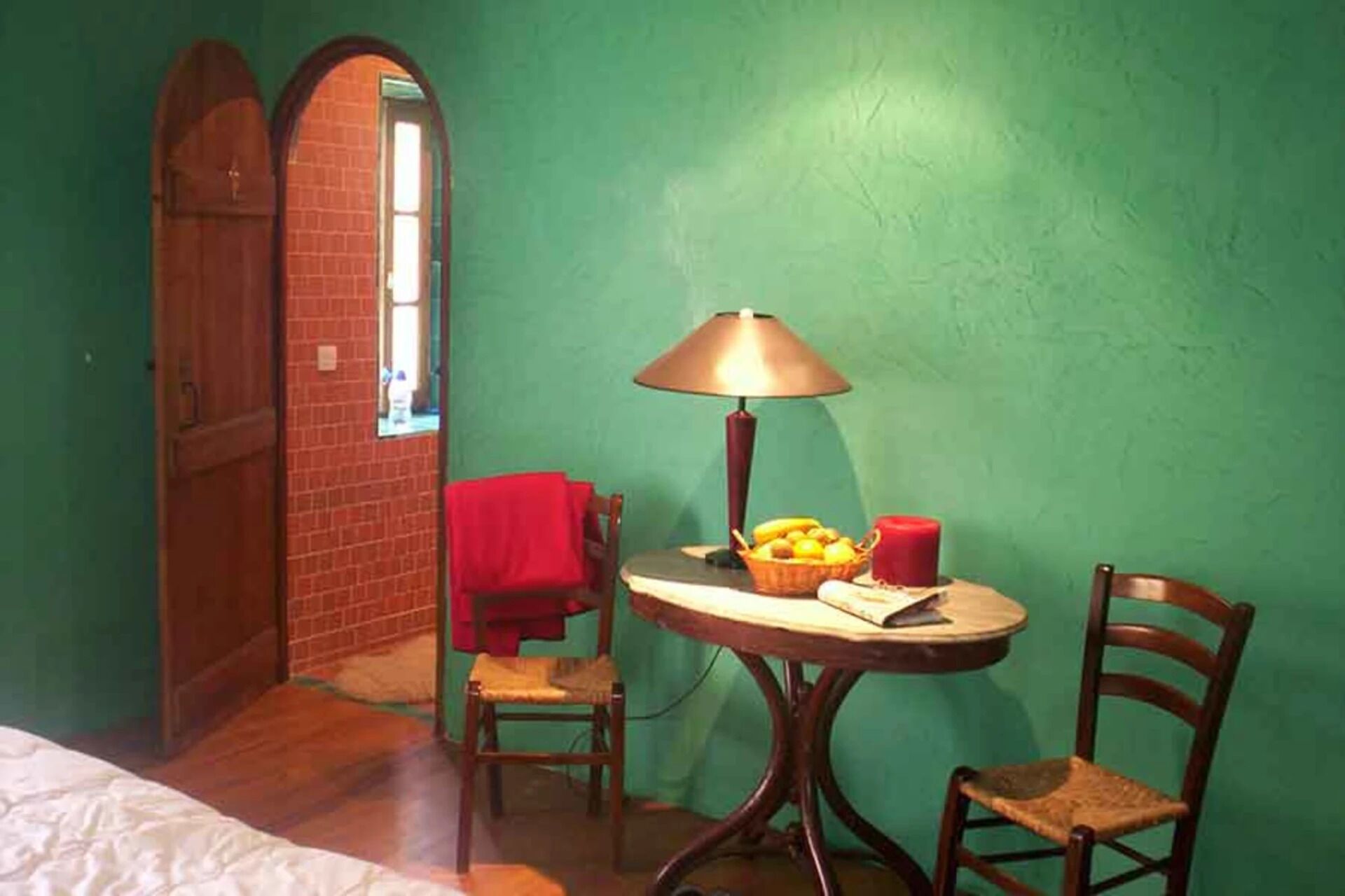 Una habitación con pared verde, puerta de madera, una mesita redonda con una lámpara, una silla y una cesta de fruta.
