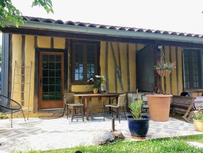 A rustic building with a tiled roof, a wooden door, and a window with a view of greenery. There is a table with chairs and a potted plant. - Domaine à Marmande (Berdoues)