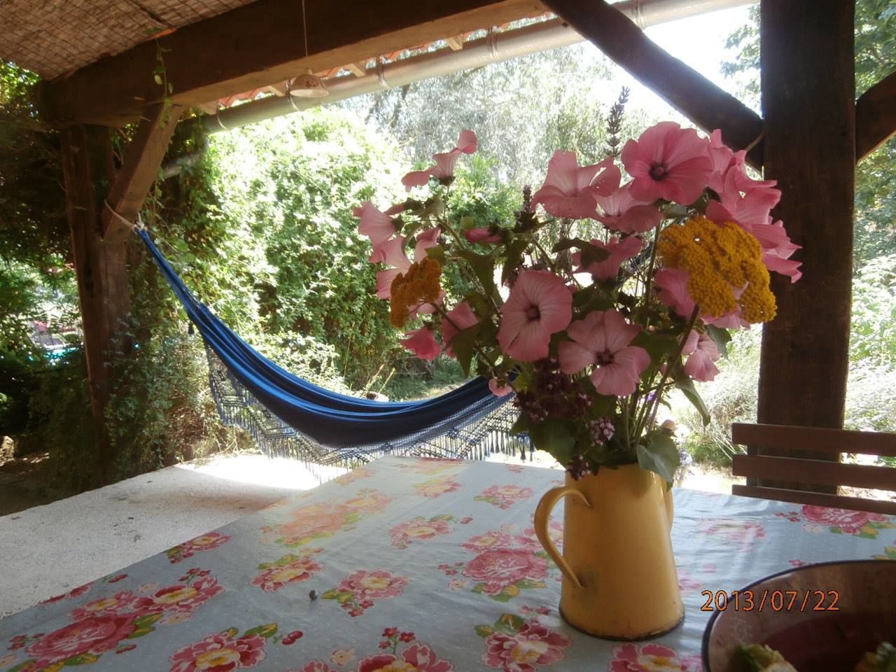 A table with a floral tablecloth, a yellow vase with flowers, and a hammock in a covered outdoor area.