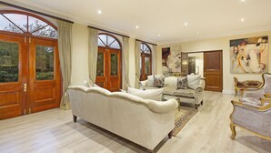 Un salon spacieux avec un grand canapé blanc, un canapé plus petit et une chaise longue. On y trouve des portes en bois avec des panneaux de verre et une grande fenêtre avec des rideaux.
