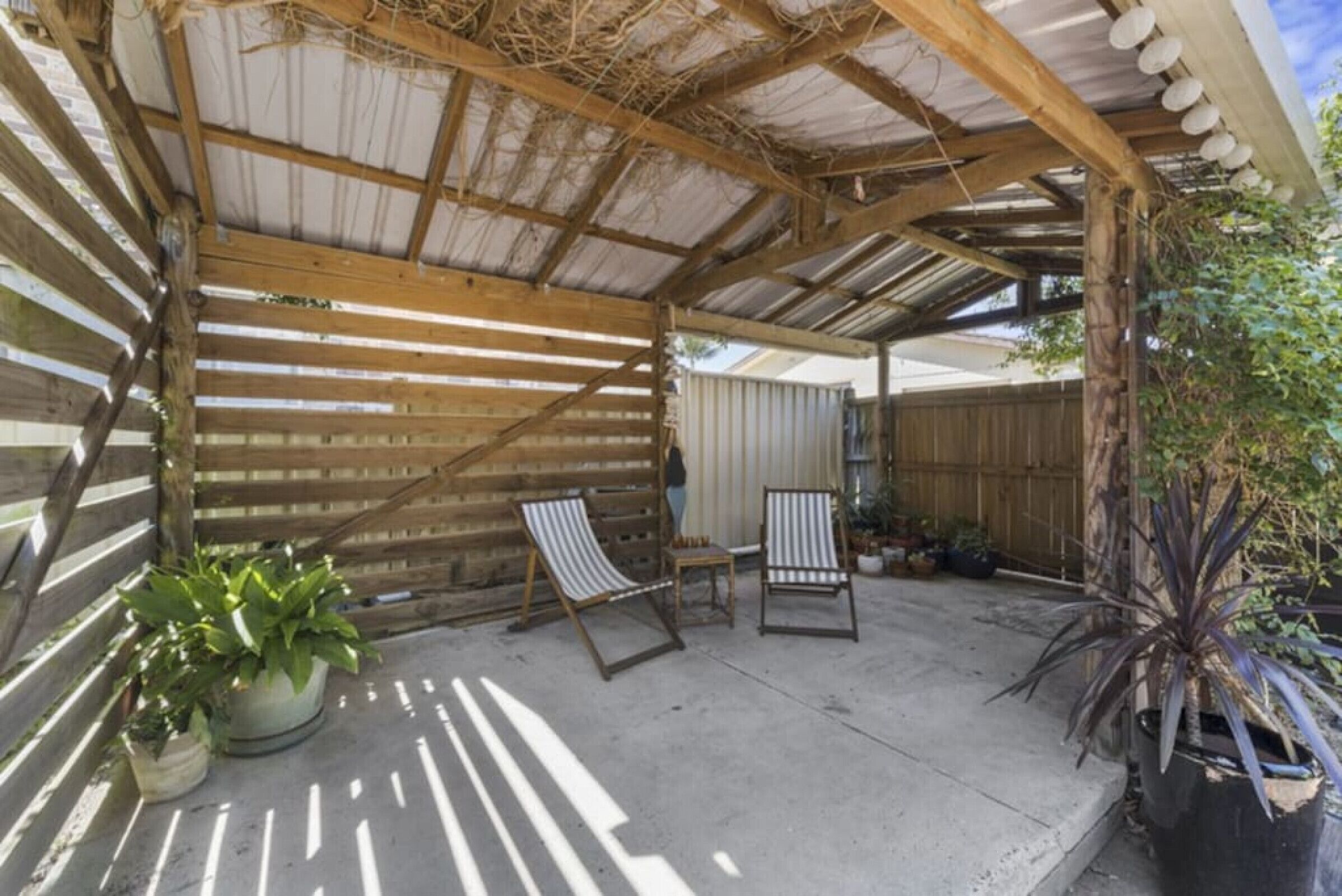 A covered patio with two striped lounge chairs, potted plants, and a wooden fence in the background.