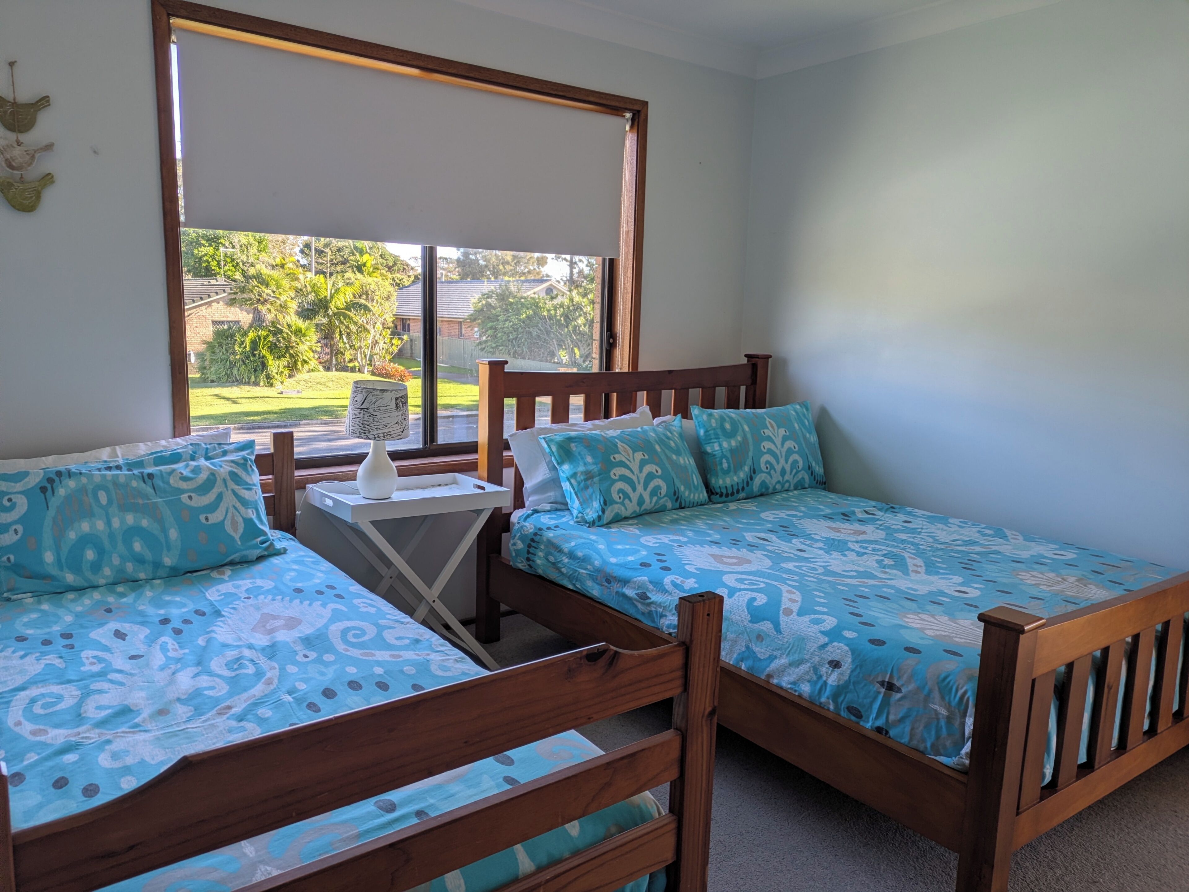 A bedroom with two wooden beds, a small bedside table with a lamp, and a window with a view of greenery and buildings.