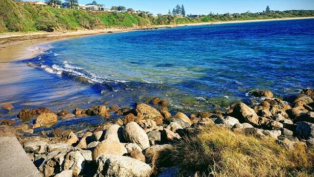 A coastal landscape with a clear blue sea, rocky shoreline, and a distant building on a hill. - 3 Min walk to the beach,  this little slice of heaven is your perfect getaway! (Culburra Beach)