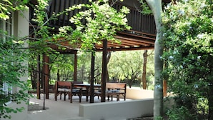 A covered outdoor seating area with wooden benches and a table, surrounded by trees and a building. - Treetops Holiday Home (Nkomazi)
