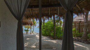 Une vue sur la plage, aperçue à travers une fenêtre ornée de rideaux, avec des palmiers et une structure dotée d’un toit de chaume.