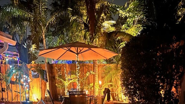 Un espace piscine extérieur avec une table et des chaises sous une pergola, entouré de plantes tropicales et d’une clôture en bois.