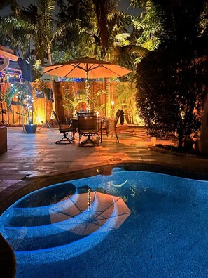 Un espace piscine extérieur avec une table et des chaises sous une pergola, entouré de plantes tropicales et d’une clôture en bois.