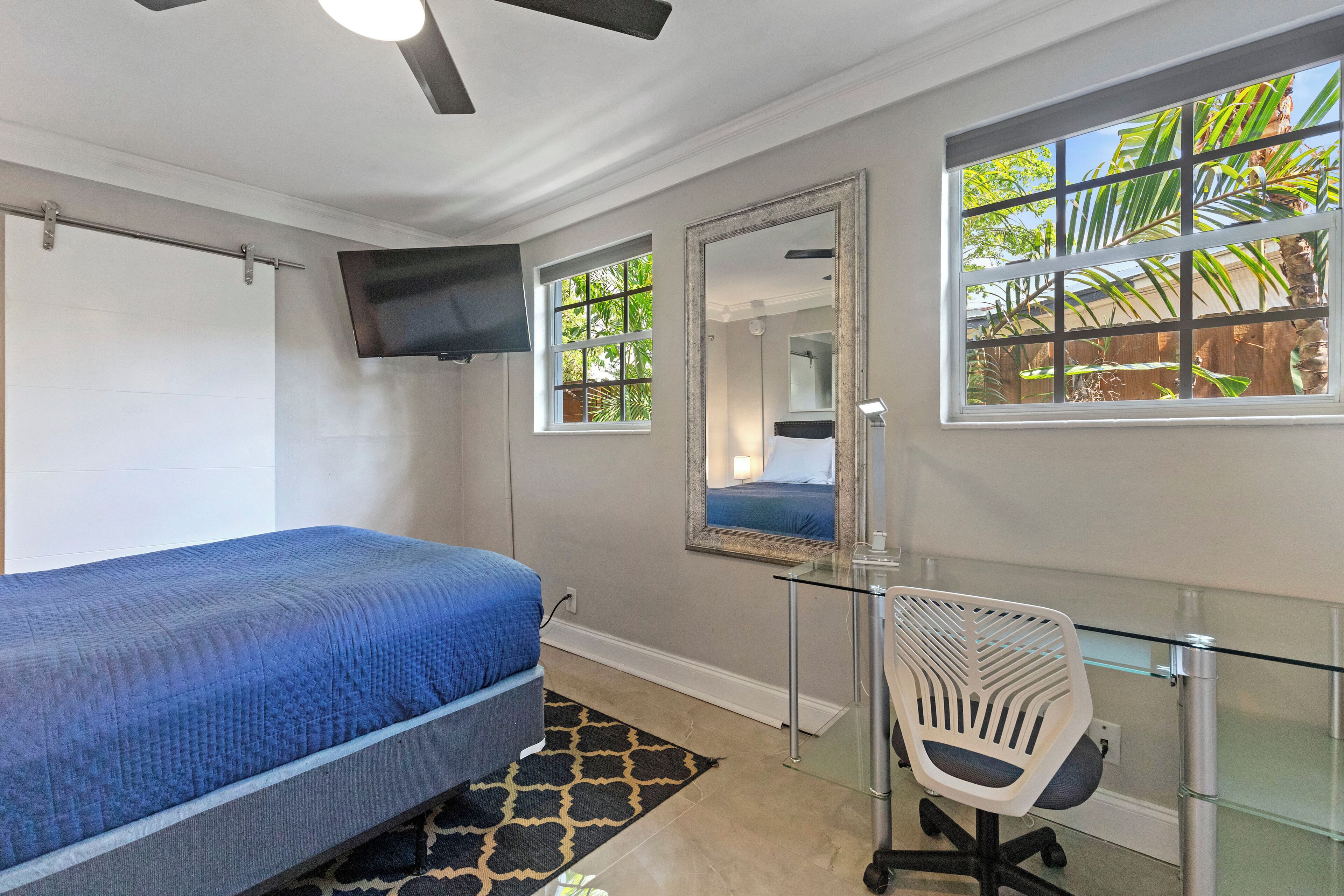 Un dormitorio con cama, escritorio, silla, televisor y ventanas que dan a palmeras.