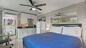 Une chambre moderne avec un grand lit, un ventilateur de plafond et un coin cuisine visible à travers une porte ouverte.