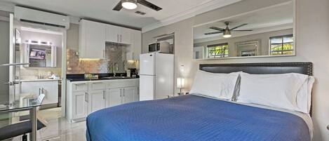 A modern bedroom with a large bed, a ceiling fan, and a kitchen area visible through an open door.