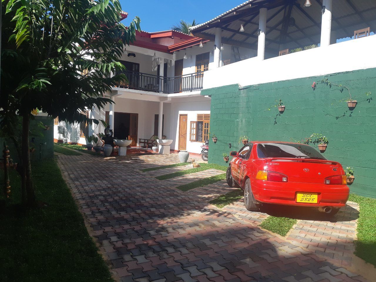 Un'auto rossa parcheggiata in un cortile con un vialetto in mattoni, circondata da vegetazione e un edificio bianco con tetto rosso.