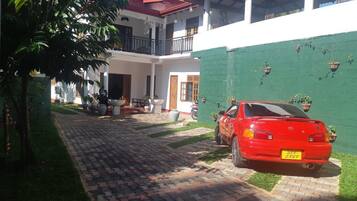 Un'auto rossa parcheggiata in un cortile con un vialetto in mattoni, circondata da vegetazione e un edificio bianco con tetto rosso.