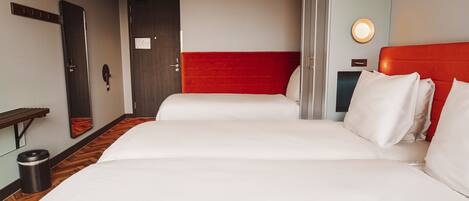 A hotel room with a large bed, a red headboard, a bedside table, a trash bin, and a wall-mounted light.