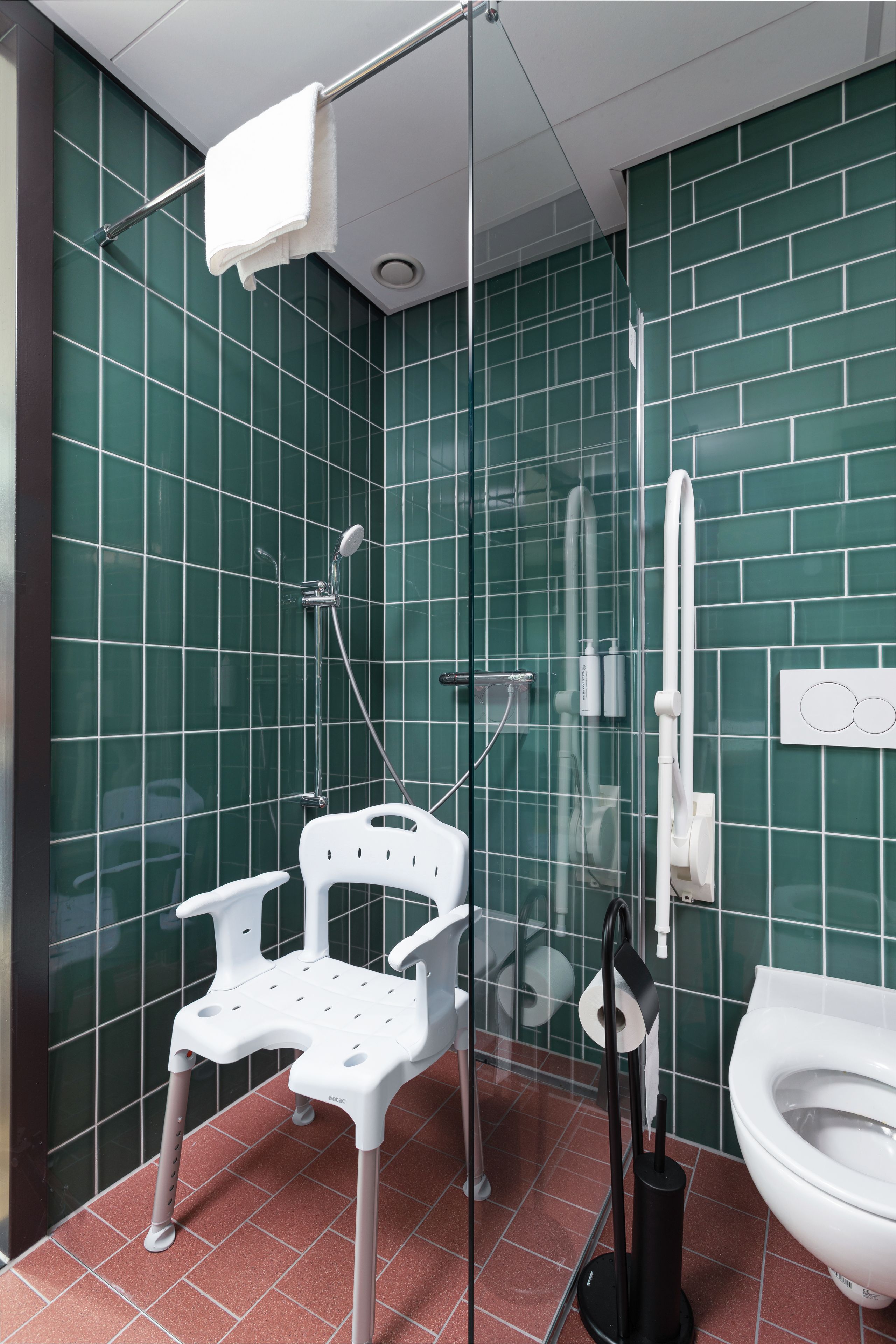Un baño con paredes de azulejos verdes, un inodoro blanco, cabina de ducha con mampara de vidrio y una silla de plástico blanca.