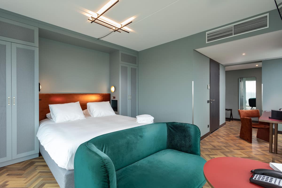 A hotel room with a bed, a green armchair, a red table, and a desk with a chair.